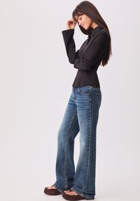 Chemise noire structurée à manches longues avec un col haut, associée à un jean large bleu délavé, présentant des coutures contrastantes et des chaussures brunes.