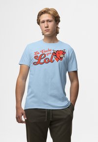 Svetle modré bavlněné tričko s červenou grafikou usmívající se krávy a textem "La Vache qui Lol" v tučném kurzívě, krátké rukávy a kulatý výstřih.