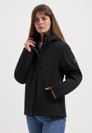 Vrouw die de kraag van een zwarte ritsjack met hoge nek en zijzak aanpast, gekleed in blauwe jeans voor een neutrale achtergrond.