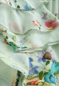 Un tessuto floreale verde chiaro presenta volant testurizzati, stampe intricate di fiori blu e rosa e cuciture delicate lungo i bordi.
