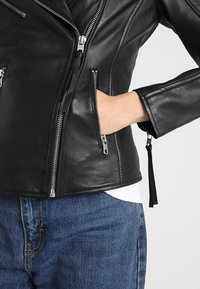 Black leather motorcycle jacket featuring silver zippers, fitted design, and accent stitching; paired with blue denim pants underneath.