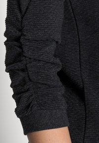 Tmavě šedý látkový návlek s texturovaným žebrovaným vzorem. Na rukávu jsou nabráné detaily a u zápěstí je přiléhavý lem.