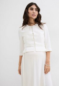 Blusa bianca a peplo con maniche a tre quarti, chiusura frontale con bottoni e tessuto liscio; sotto si trova un vestito bianco fluido.