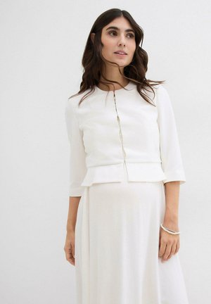 Blusa bianca a peplo con maniche a tre quarti, chiusura frontale con bottoni e tessuto liscio; sotto si trova un vestito bianco fluido.