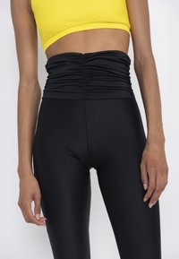 Black, high-waisted leggings with gathered texture at the waist. Paired with a sleeveless, bright yellow cropped top. Smooth, stretchy fabric.