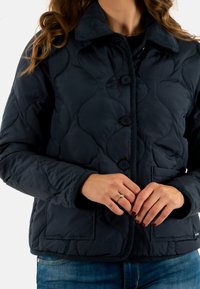 Giacca imbottita blu navy con chiusura frontale a bottoni, colletto in cotone a coste, due tasche laterali e polsini elasticizzati; texture morbida e imbottita.
