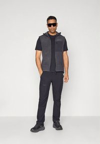 Colete preto de lã com bolsos com fecho, combinado com uma t-shirt preta e calças a condizer. Usa óculos de sol e sapatos desportivos pretos.