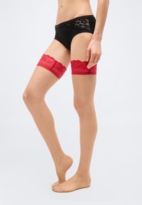 Κάτω μέρος σώματος ενός ατόμου που φοράει μαύρα εσώρουχα με δαντέλα και διάφανα κόκκινα καλσόν δαντέλας πάνω από το γόνατο, που στέκεται στις μύτες των ποδιών πάνω σε μονόχρωμο φόντο.