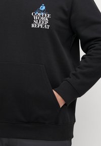 Schwarzer Kapuzenpullover mit einer Vordertasche, versehen mit weißem und blauem Text mit der Aufschrift "KAFFEE ARBEIT SCHLAF WIEDERHOLEN" und einer grafischen Illustration.