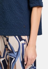 Granatowa teksturowana bluzka z wzorzystym dołem w niebieskie, beżowe i brązowe linie; dłoń spoczywa na tkaninie, prezentując warstwowe wzory.