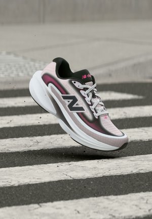 Ροζ ανοιχτό και μαύρο αθλητικό παπούτσι New Balance που προσγειώνεται σε άσφαλτο κοντά σε λευκή διάβαση πεζών με μικρά σωματίδια στον αέρα.