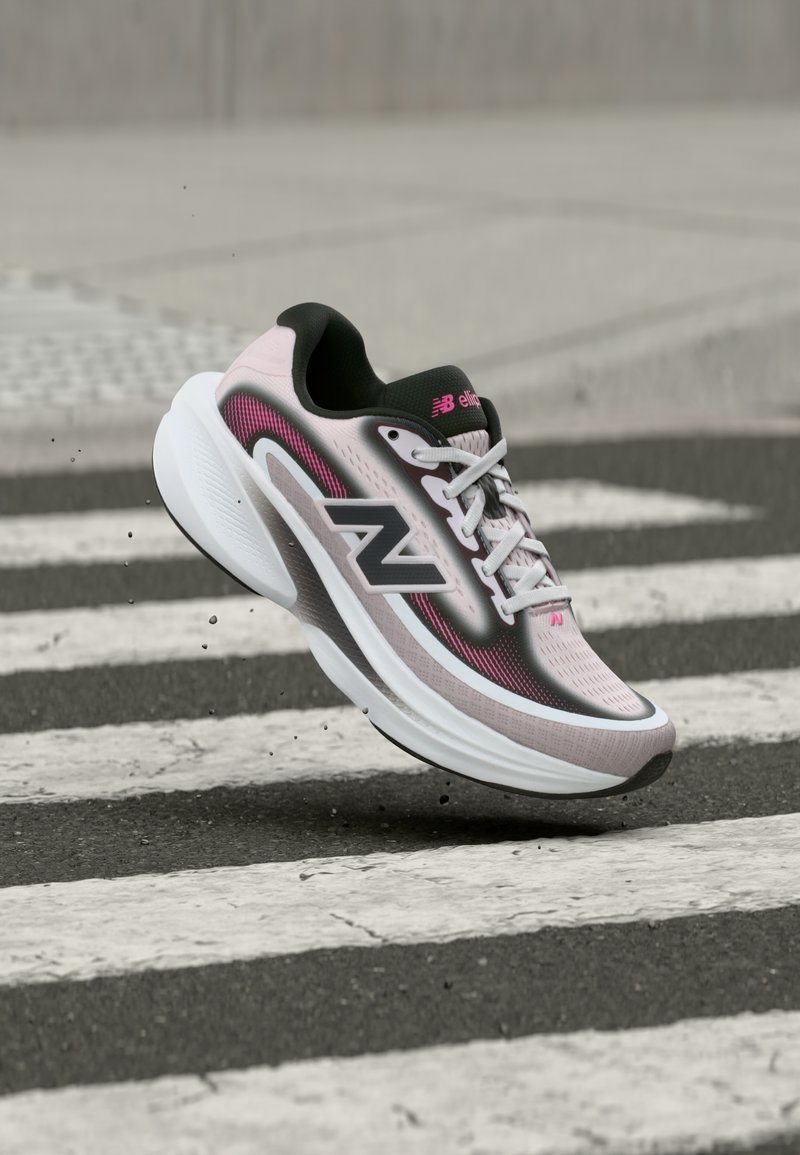 Chaussure de course New Balance rose clair et noire atterrissant sur l'asphalte près d'un passage piéton blanc avec de petits débris en suspension dans l'air.
