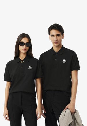 Zwei Personen tragen enganliegende schwarze Polohemden mit einem weißen Lacoste-Logo. Das Hemd hat einen Kragen und kurze Ärmel. Eine Person trägt eine Sonnenbrille.