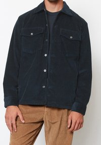 Ciemnoniebieska kurtka sztruksowa z dwoma kieszeniami na piersi, zapięciem na guziki i klasycznym kołnierzem. Noszona z beżowymi, prążkowanymi spodniami.