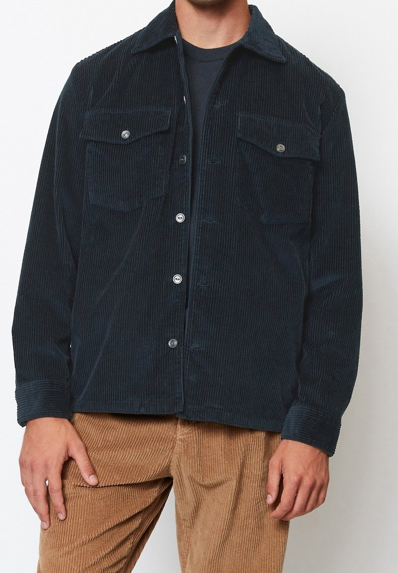 Ciemnoniebieska kurtka sztruksowa z dwoma kieszeniami na piersi, zapięciem na guziki i klasycznym kołnierzem. Noszona z beżowymi, prążkowanymi spodniami.