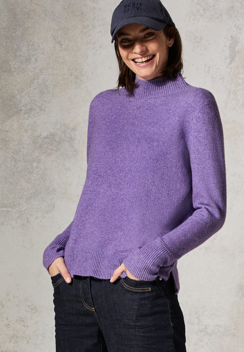 Lila gestrickter Pullover mit hohem Kragen, gerippten Bündchen und Saum. Getragen über dunklen Jeans. Strukturiertes Gewebe mit weichem Finish.