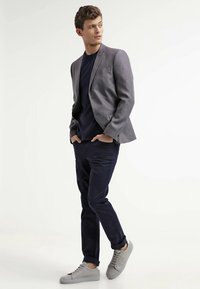 Pelēks blazers virs tumsas zila krekla, kopā ar tumši zilām biksēm un gaiši pelēkiem sporta apaviem. Apģērbs izceļas ar elegantu dizainu un piegriezta piekrišanu.