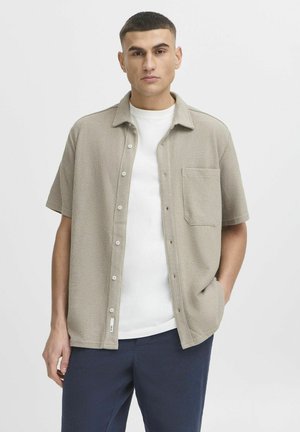 Camisa de manga corta, con botones, en tejido texturizado beige con un bolsillo en el pecho, llevada sobre una camiseta blanca, combinada con pantalones azul marino.