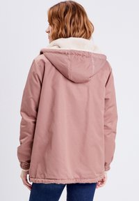 Rosa jacka med en mysig beige pälsfoderhuva, elastiska ärm- och nederkanter. Enkelt design med en något lös passform för komfort.