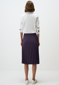Gonna plissettata viola realizzata in tessuto leggero, abbinata a una camicia bianca con colletto, vista di schiena, con sneakers bianche visibili.