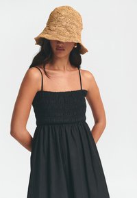 Vestido de tirantes delgados con smock negro, que presenta un corsé ajustado y una falda fluida. Combinado con un sombrero de cubo de paja texturizada.