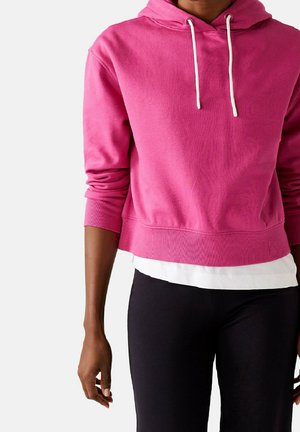 Persoon die een felroze cropped hoodie draagt met witte trekkoorden over een wit hemd en zwarte broek, staand tegen een effen achtergrond.