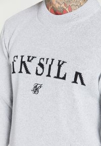 Un pull en maille gris clair avec un texte noir et un petit logo sur le devant. Encolure et bords de manches côtelés. Le texte est stylisé.