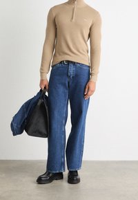 Uomo che indossa un maglione beige con zip, jeans blu e stivali neri, mentre tiene in una mano una borsa nera e una giacca di denim.