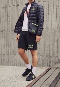 Giacca nera imbottita con collo alto, t-shirt bianca con logo, pantaloncini neri con accenti verdi e sneakers nere con dettagli verdi.