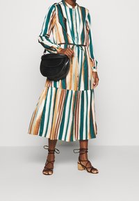 Abito midi a righe in teal, marrone e crema con chiusura a bottoni e gonna a strati. Borsa a tracolla nera e sandali marroni con cinturini.