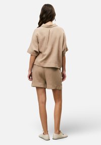 Beige textured short-sleeve top with a collar and pleat detail, paired with matching shorts. Model wears light-colored shoes.