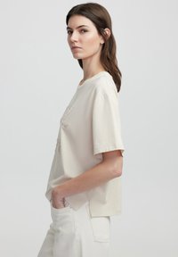 Camiseta de algodón beige con mangas cortas, cuello redondo y un ajuste ligeramente holgado. Presenta bordados decorativos en la parte delantera.