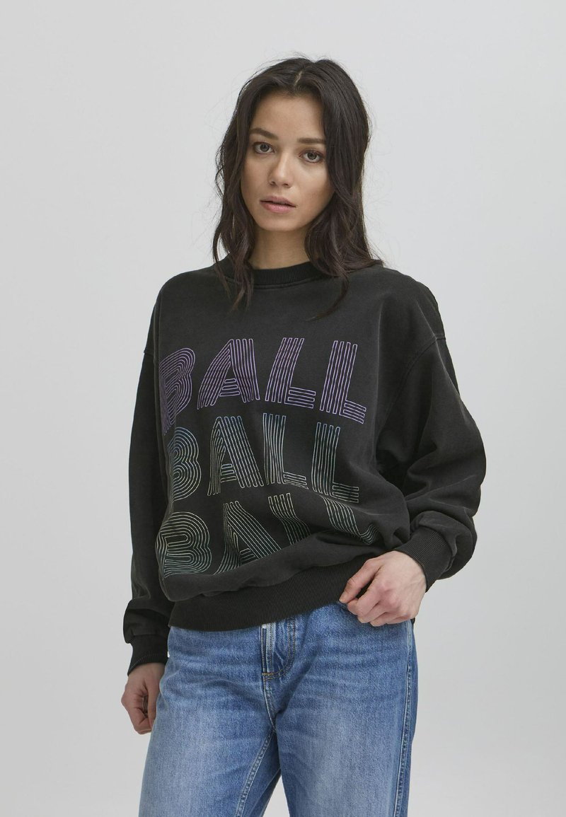 Svart hettegenser med teksten "BALL" i varierende linjestiler og farger. Rund hals, lange ermer, avslappet passform. Brukt sammen med lyseblå jeans.
