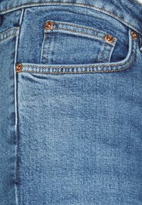 Vaqueros de mezclilla con un lavado azul claro, detalles de herrajes de cobre y un pequeño bolsillo frontal. La tela presenta un acabado suave y texturizado.