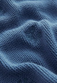 Modrý pletený materiál s texturovaným tkaným vzorem. Obsahuje měkké, pružné vlákna a jemnou květinovou výšivku ve slabě tmavší modré.