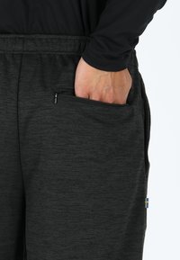 Zwarte sportbroek met een ritszak op de heup, gemaakt van een textuurstof en met een kleine blauwe en gele accent nabij de zak.