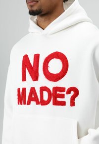 Witte hoodie met rood, gestructureerd borduursel met de tekst "NO MADE?" duidelijk zichtbaar. Voorzien van een capuchon met trekkoord en een kangaroo-zak.