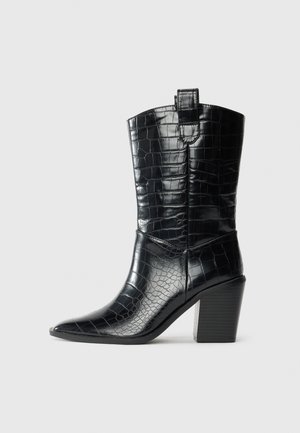 Bota ocidental preta até o meio da panturrilha com bico apontado, salto bloco e textura de couro brilhante em relevo de crocodilo em fundo branco.