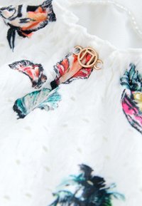 Tela blanca texturizada con estampados florales coloridos, con un cierre circular dorado en el escote. Detalle delicado y diseño en capas.