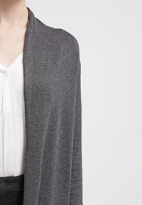 Cárdigan gris hecho de una tela suave, diseño de frente abierto, mangas largas y un ajuste relajado, superpuesto sobre una blusa blanca. Textura suave.