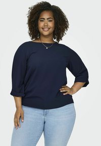 Blusa blu navy con maniche a 3/4 e risvolti, caratterizzata da un collo rotondo e tessuto morbido, abbinata a jeans azzurri.
