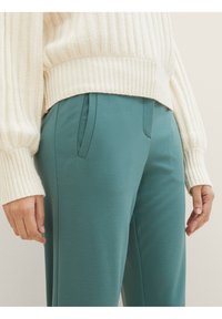 Lichte crème geribbelde gebreide trui gecombineerd met smaragdgroene, stretchy, op maat gemaakte broeken met zijzakken en een soepele textuur.