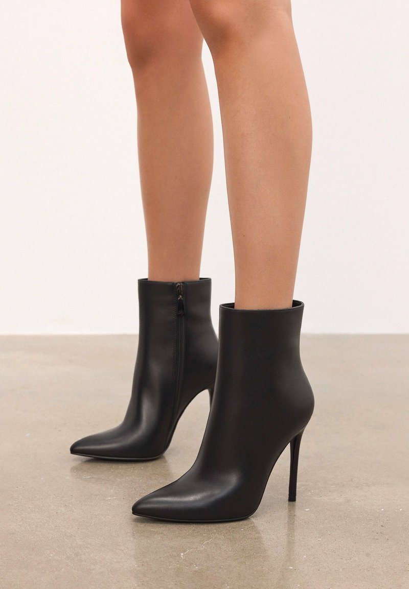 Botines de cuero negro con punta afilada, tacón alto de aguja y cierre lateral con cremallera. Textura suave y diseño elegante.