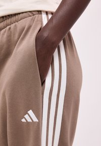 Personne à la peau foncée avec la main dans la poche d'un pantalon de survêtement beige avec trois bandes blanches et un petit logo blanc sur la cuisse.