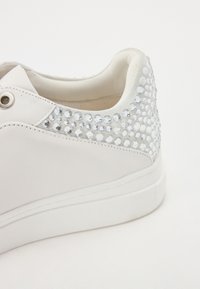 Valge nahast tennisetoss, millel on lame kummist sisetald ja mis on kaunistatud seljaosa selgete gemstone'idega, et lisada detaili.