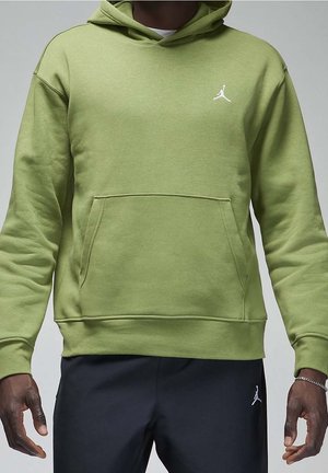 Mężczyzna w jasnozielonej bluzie z kapturem i czarnych spodniach, oba z małymi białymi logo Jumpman, stoi z opuszczonymi ramionami na szarym tle.