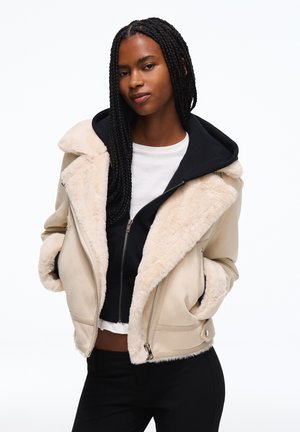 Jeune femme aux longs cheveux tressés porte une veste en shearling beige sur un sweat à capuche zippé noir et une chemise blanche, posant les mains dans les poches.