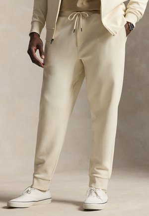 Mand iført beige joggingbukser med snøre, cremefarvet jakke og hvide sneakers, stående med den ene hånd i lommen og armbåndsur synligt.