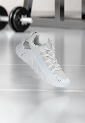 Baskets athlétiques gris clair et blanc avec une semelle épaisse et des lacets, flottant au-dessus d'un sol de gym réfléchissant avec des poids flous en arrière-plan.