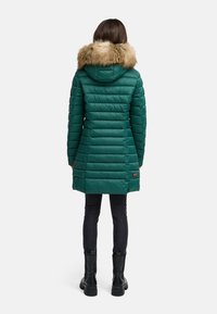 Parka verde acolchada con capucha forrada de piel sintética, patrón de acolchado horizontal, cierre con cremallera y bolsillos laterales, combinada con jeans ajustados negros y botas.